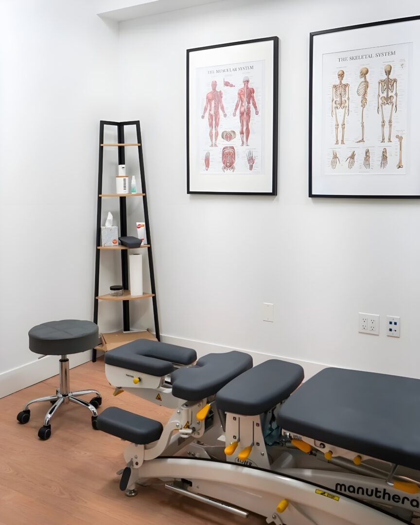 A modern chiropractic treatment room featuring a Manuthera adjustment table, a rolling stool, and anatomical charts of the skeletal and muscular systems on the wall.
