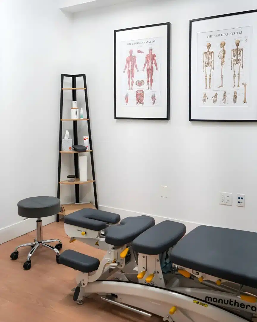A modern chiropractic treatment room featuring a Manuthera adjustment table, a rolling stool, and anatomical charts of the skeletal and muscular systems on the wall.