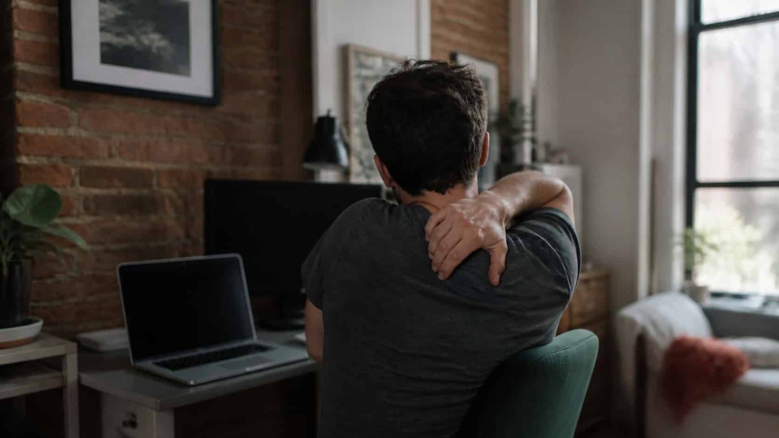 Person at a Brooklyn home office desk reaching for pain between your shoulder blades, treated at Brooklyn Chiropractic Care in Greenpoint